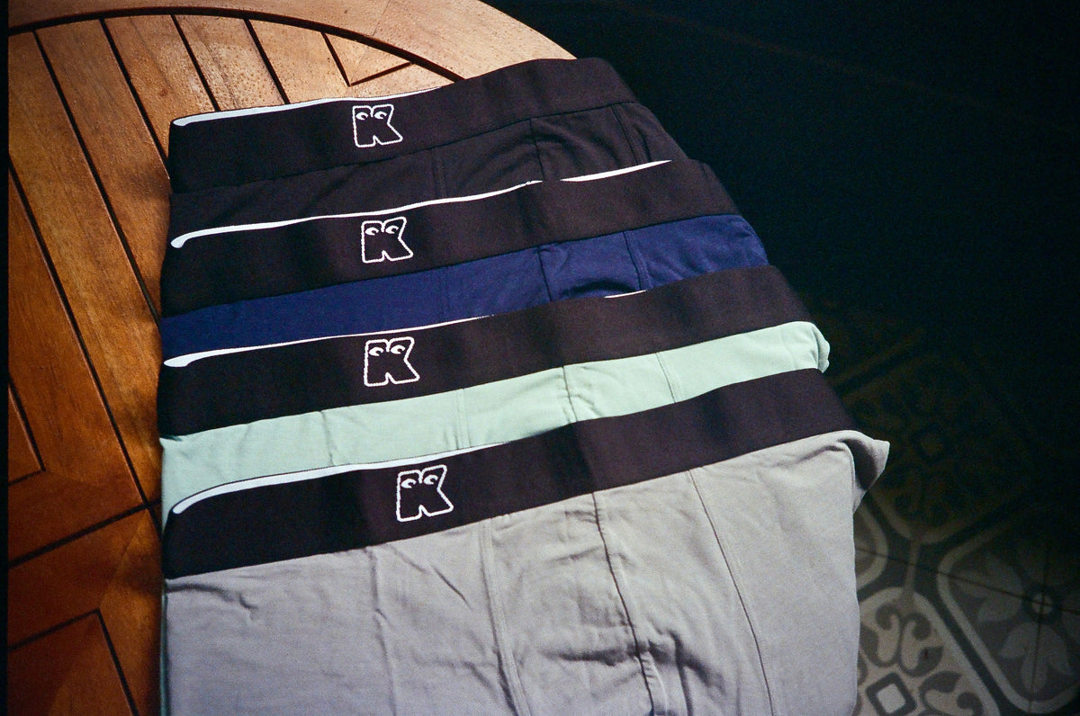 Four folded Kokoruno underwear pairs on a wooden table, with black waistbands and embroidered logos.