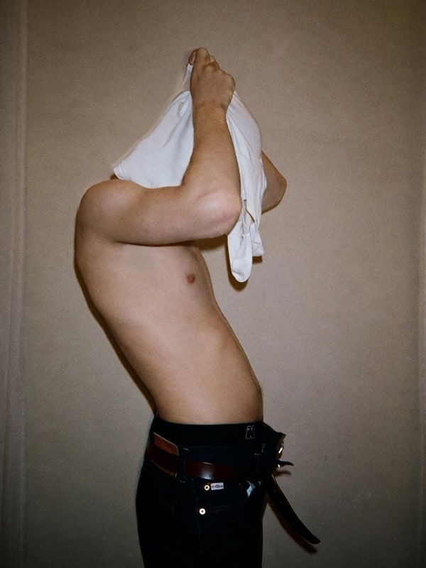 Person with tattoos lifting a white shirt to reveal their chest against a plain background