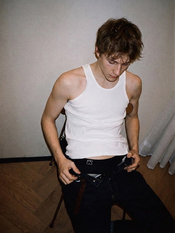 Man wearing a white tank top and black pants with tattoos on his arms, standing against a plain wall.